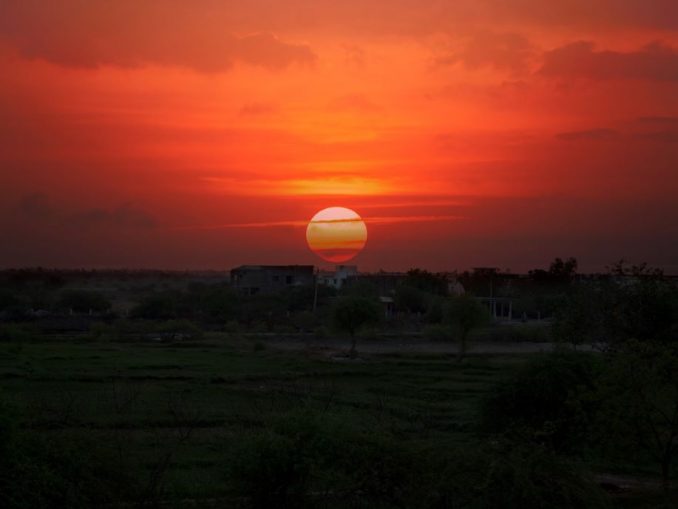 Coucher de soleil en phase terminale (dernier appel) à la campagne, verdure dans la pénombre à l’avant-plan et ciel rougeâtre avec nuages en arrière-plan.
