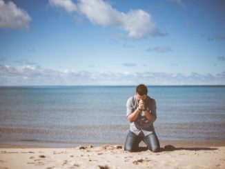 Homme agenouillé (la prière) sur la plage à l’avant-plan; mer bleue et calme à l’arrière-plan, sous un ciel bleu avec nuages à l’horizon et cumulus en altitude.