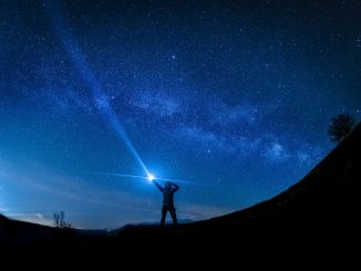 Silhouette d’un homme pointant une lampe de poche vers le ciel à l’avant-plan. Ciel bleu étoilé à l’arrière-plan.