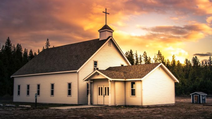 Petite église en bois brun et blanc à l’avant-plan. Forêt de conifères à l’arrière-plan sous un ciel partiellement nuageux durant le jour.