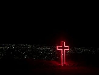 Croix de néon rouge à l’avant-plan. Milliers de points lumineux d’une ville sous un ciel complètement noir à l’arrière-plan.