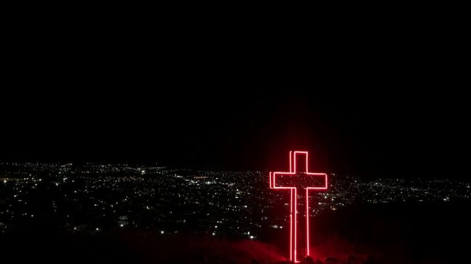 Croix de néon rouge à l’avant-plan. Milliers de points lumineux d’une ville sous un ciel complètement noir à l’arrière-plan.