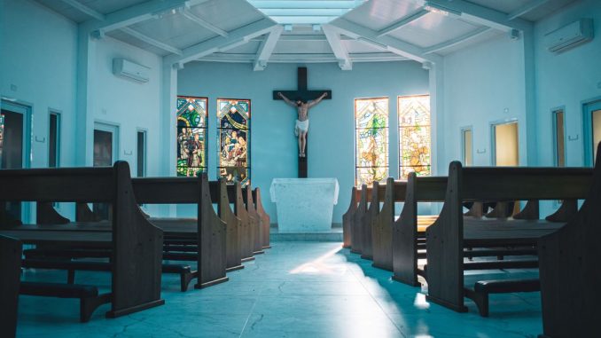 Bancs d’église en bois brun foncé à l’avant-plan. Au fond, crucifix au centre et deux vitraux de chaque côté. Murs, plafond et plancher de couleur bleu ciel.