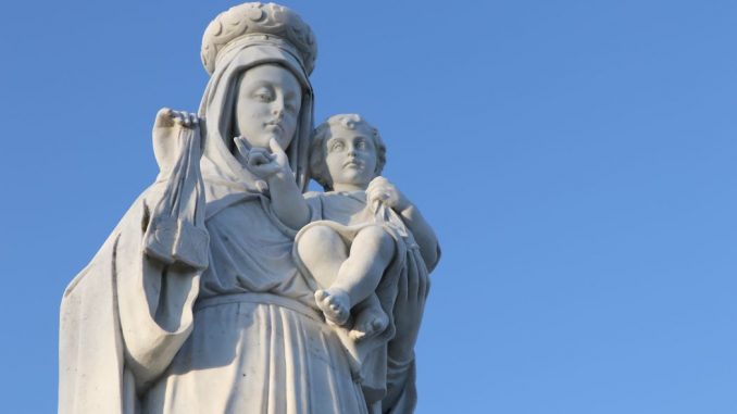 Statue de la Vierge Marie avec l’enfant Jésus dans ses bras, tous les deux habillés en blanc. Ciel bleu.