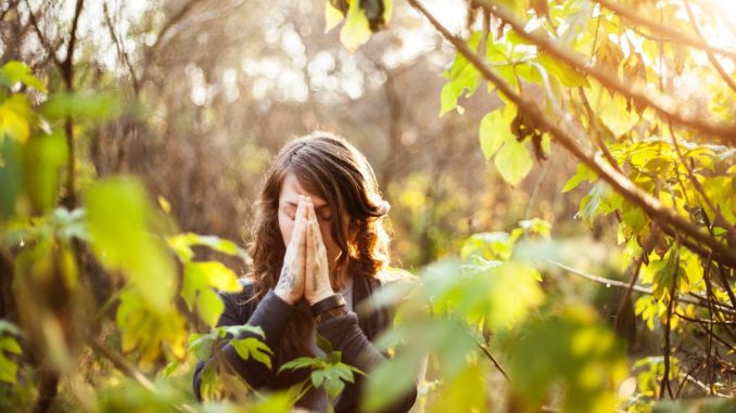 Femme en veste noire, mains jointes devant le visage, debout près d’un arbre à feuilles jaunes durant le jour.