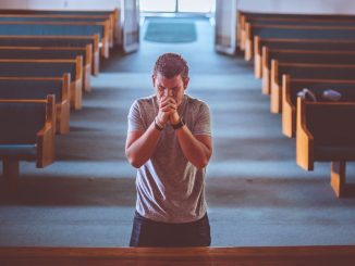 Homme en prière, avec les mains jointes devant la bouche, debout devant l’autel d’une église ou d’une chapelle.