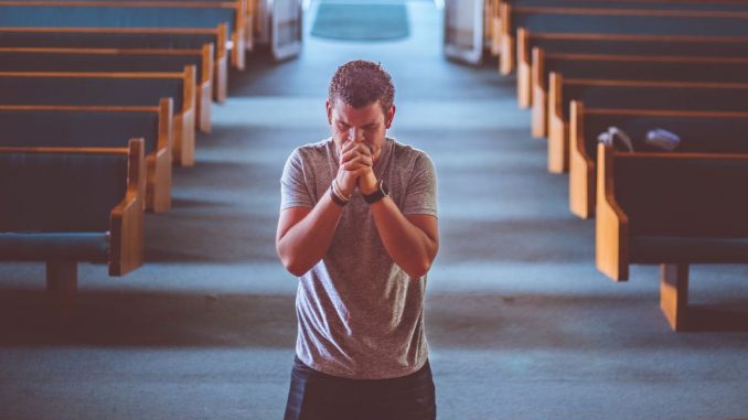 Homme en prière, avec les mains jointes devant la bouche, debout devant l’autel d’une église ou d’une chapelle.