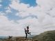 Homme soulevant une femme au sommet d’une montagne durant le jour. Ciel parsemé de gros nuages blancs.