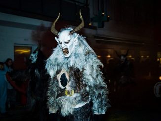 Personne portant un crâne cornu et une fourrure d’animal blanc et noir. Personnages en arrière-plan dans la pénombre.