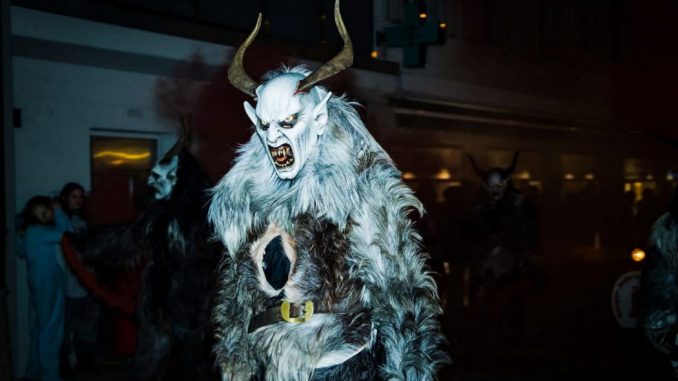 Personne portant un crâne cornu et une fourrure d’animal blanc et noir. Personnages en arrière-plan dans la pénombre.