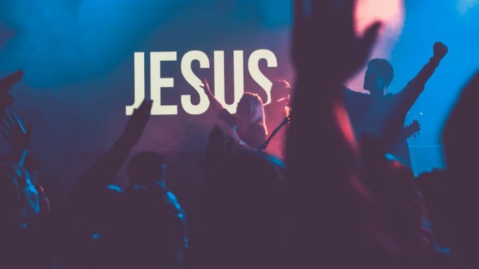 Concert religieux joué par un groupe sur scène. Jésus, affiché en très gros caractères blancs, apparaît au fond de la scène faiblement éclairée.