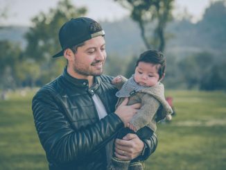 Homme souriant, portant une veste en cuir noire, tenant un bébé par son bras gauche. Image floue en arrière-plan.