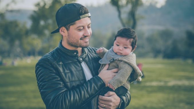 Homme souriant, portant une veste en cuir noire, tenant un bébé par son bras gauche. Image floue en arrière-plan.