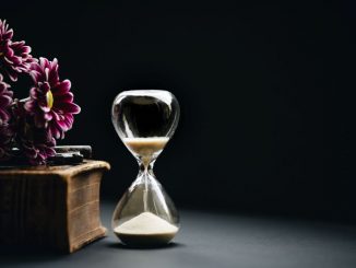 Sablier clair à côté de fleurs roses posées sur un coffret brun.