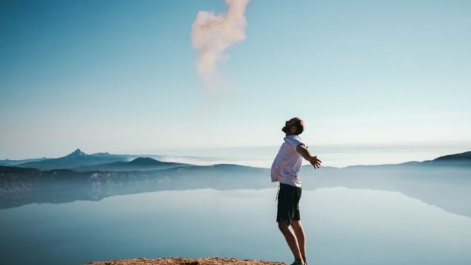 Homme debout sur le sable les bras en croix à côté d’un plan d’eau calme.