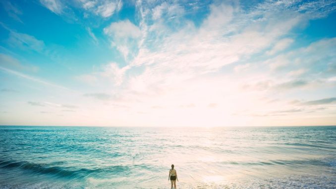 Femme debout dans la mer devant le soleil voilé par des nuages blancs.