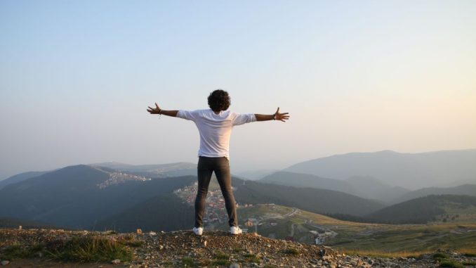 Homme en t-shirt blanc et pantalon gris debout au sommet d’une montagne.