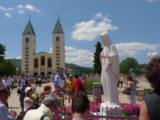 Many pilgrims (convert the whole world) on the square of Saint Peter's church in Medjugorje. In the foreground is a statue of the Virgin Mary. In the background, the facade of the church and the mountain under a blue sky with cumulus clouds.