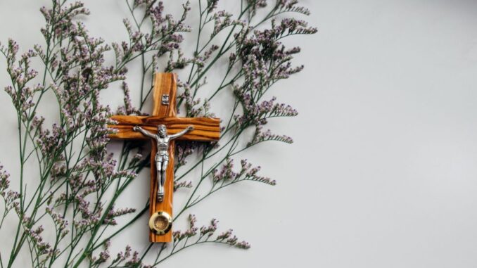 A brown wooden crucifix (take the cross) on a gray wall with flowers.