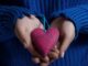 Close-up on a red fabric heart offered in the palm of the right hand (give me your heart), which rests on the palm of the other hand, by a person in a blue wollen sweater.