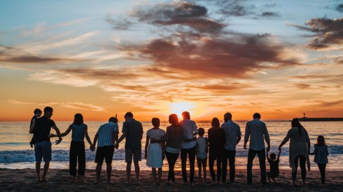 Large family united by love (love in your families), standing side by side on the beach, facing the sea and the setting sun on the horizon. Blue sky with red tinted clouds near the sun.
