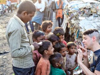 On the edge of an alley, a group of Indian children accompanied by an adult listening with joy to a stranger (apostle of love) in the foreground. Litter on the right and other people not quite visible are watching the scene in the background.