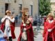 Three persons in costume (red coat, hat and fur collar) walk side by side in broad daylight behind a car to carry the cross in the foreground. The person in the center carries the cross. A building is on the left and an icon of the Virgin on the right. Several people circulate behind but in the opposite direction of the procession In the background a tree and another building.