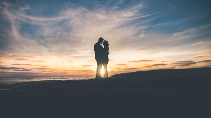 Couple embracing on a beach (live love in your life) in the semi-darkness in the foreground but lit by the reddish sky of the setting sun on the horizon in the background.