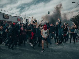 Angry protesters (peaceless world), hands and fists in the air in the foreground. The roof of a small commercial building is seen above protesters on the left and shrubs on the right. Black smoke in the background under a sky with white and darker clouds.