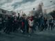 Angry protesters (peaceless world), hands and fists in the air in the foreground. The roof of a small commercial building is seen above protesters on the left and shrubs on the right. Black smoke in the background under a sky with white and darker clouds.