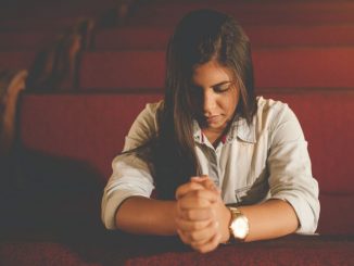 Praying woman (pray for plan of salvation) kneeling in a dark brown church pew in the foreground. Soft lighting. We can see three other pews behind.