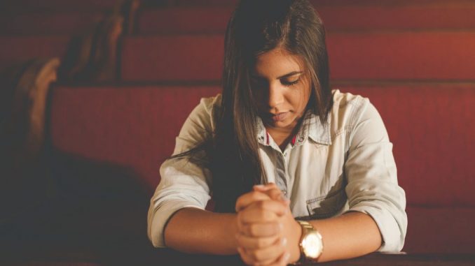 Praying woman (pray for plan of salvation) kneeling in a dark brown church pew in the foreground. Soft lighting. We can see three other pews behind.