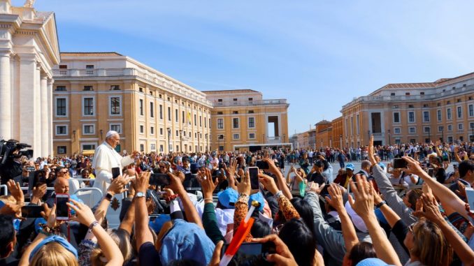 Crowd of people greeting the Pope (joy of faith) in a large square in front of a cathedral on the left side. Pink-colored buildings around the square, under a blue sky partially obscured by a thin layer of white clouds.