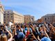 Crowd of people greeting the Pope (joy of faith) in a large square in front of a cathedral on the left side. Pink-colored buildings around the square, under a blue sky partially obscured by a thin layer of white clouds.