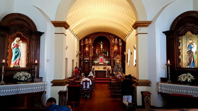 People praying in a small church (live your faith). In the foreground, the altar of the Virgin on the left and the altar of Saint Joseph on the right. High altar at the back. A few people are seated and others are on their knees. The benches and the floor are dark brown wood.
