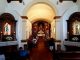 People praying in a small church (live your faith). In the foreground, the altar of the Virgin on the left and the altar of Saint Joseph on the right. High altar at the back. A few people are seated and others are on their knees. The benches and the floor are dark brown wood.