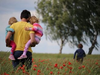 A father with a child in each arm (your life is a gift for others) and a mother walk in red flower field. Two leafy trees further on the right. Blue sky tinted with a thin layer of white clouds in the background.