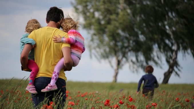 A father with a child in each arm (your life is a gift for others) and a mother walk in red flower field. Two leafy trees further on the right. Blue sky tinted with a thin layer of white clouds in the background.