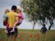 A father with a child in each arm (your life is a gift for others) and a mother walk in red flower field. Two leafy trees further on the right. Blue sky tinted with a thin layer of white clouds in the background.