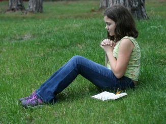 Young girl sitting on the grass, hands folded, eyes closed, and face beaming with joyce and peace. Beside her a bible. In the background, we see the base of some trees.