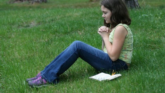 Young girl sitting on the grass, hands folded, eyes closed, and face beaming with joyce and peace. Beside her a bible. In the background, we see the base of some trees.