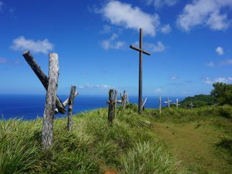 Brown wooden cross near a path (path to salvation), on green grass field along the sea, under blue sky during day time.