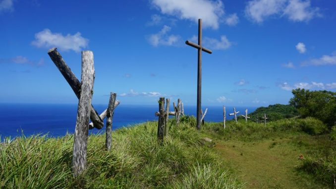 Brown wooden cross near a path (path to salvation), on green grass field along the sea, under blue sky during day time.