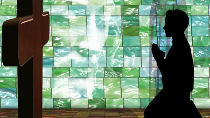 Profile silhouette of a person kneeling on a ceramic floor in front of a small brown wooden cross. In the background, a wall made of glass mosaic tiles in different colors of green and white. You can see the features of a face close-up.