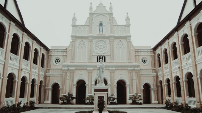 Square (way of holiness) in front of a beige church inserted between two buildings. White statue in the center. Cloudy sky.