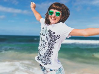 Smiling woman with crossed arms, wearing white and black shirt, jumpiing near seashore under blue sky.