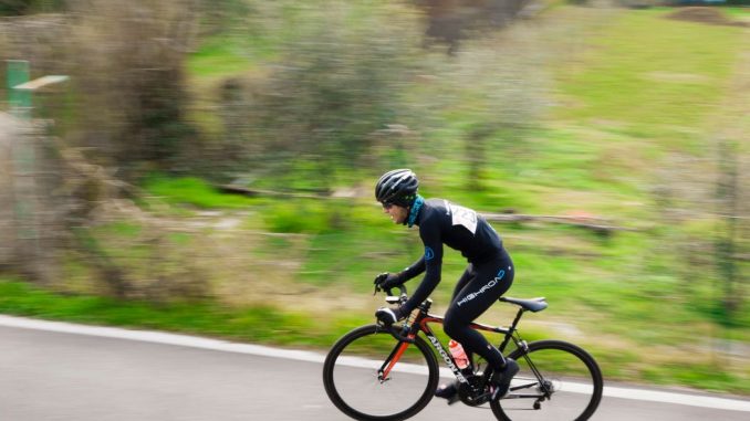Cyclist dressed in black climbing a moutain road (accept pain) during daytime. Blurred landscape in the background.