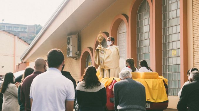 Priest presenting the monstrance to a group of people (know my Son) in front of a brown building during daytime.