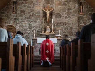 People in adoration inside a church, on either side of the central aisle. Priest kneeling before the monstrance on the altar.