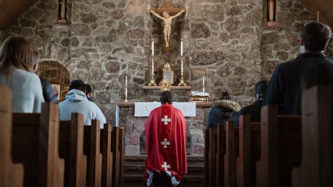 People in adoration inside a church, on either side of the central aisle. Priest kneeling before the monstrance on the altar.